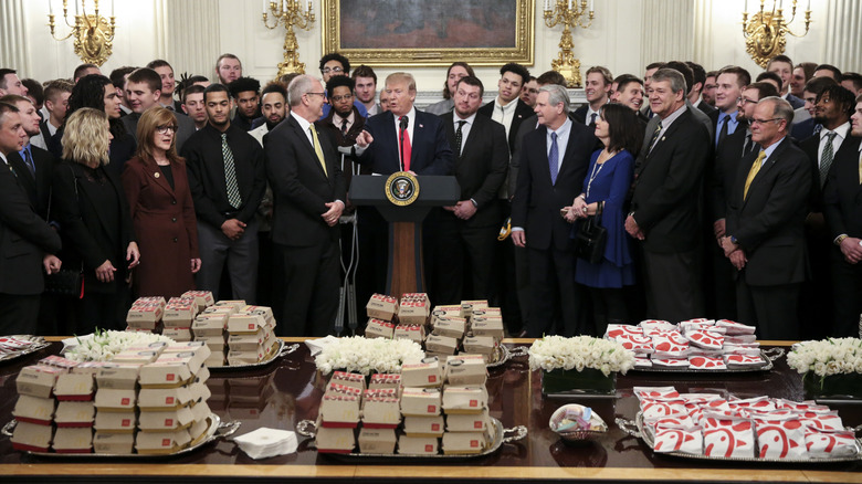 Table full of fast food burgers and sandwiches in front of Donald Trump surrounded by officials and a college football team