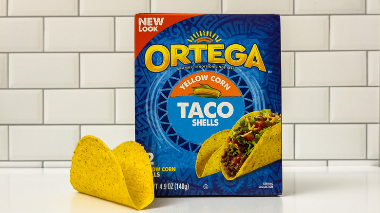 A box of Ortega's yellow corn taco shells, with a shell learning on the left side of the box