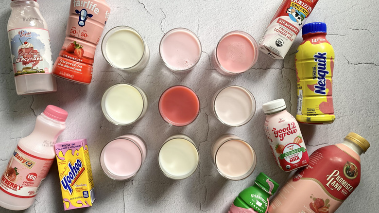 A variety of strawberry milks in individual glasses, surrounded by strawberry milk bottles and cartons from different brands
