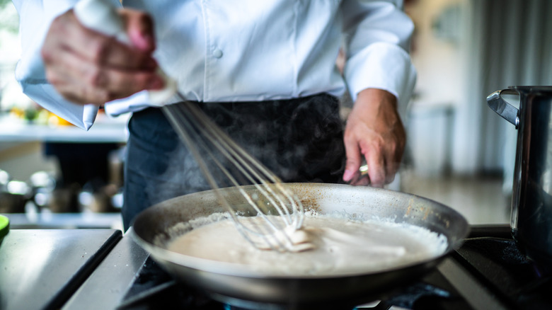 A chef whisking a thick white sauce in a shallow stainless steel saucepan