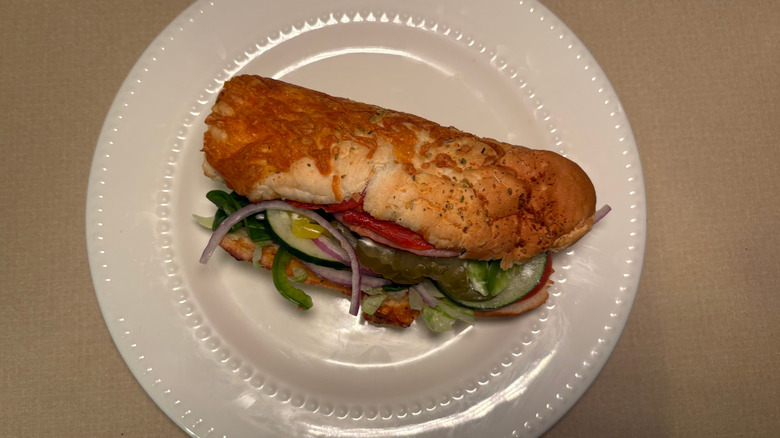 A sub on Italian herbs and cheese bread with a variety of vegetables and meats on a plate.
