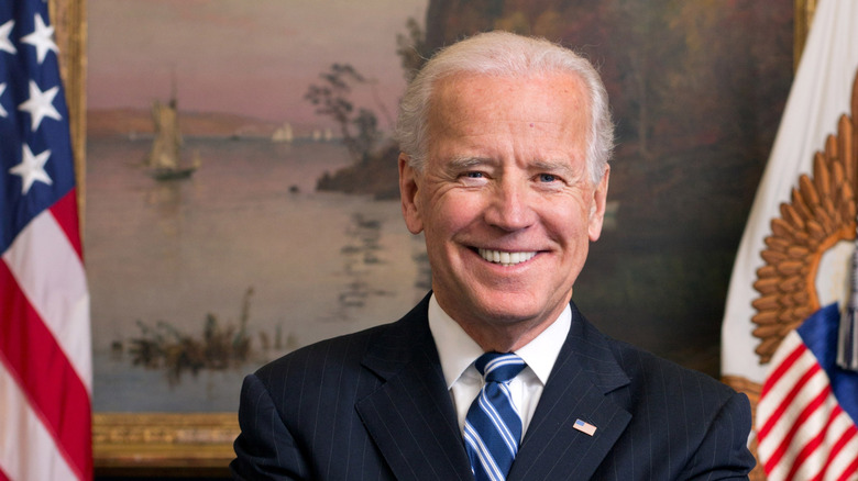 President Joe Biden in front of painting, with two flags behind him, one on each side
