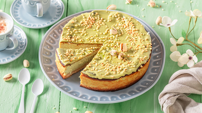 a whole pistachio cheesecake on a light green wooden table with two cups of coffee