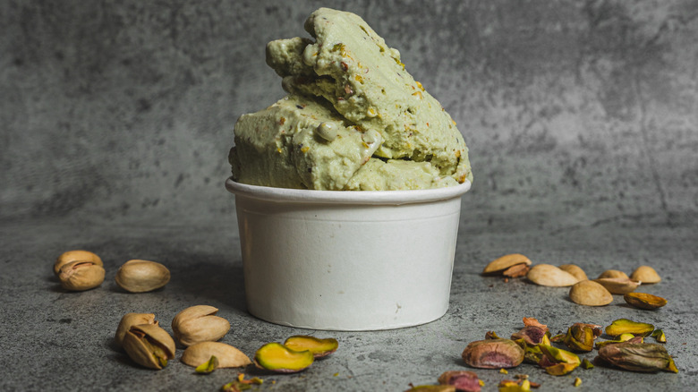 pistachio gelato in a white cup on a gray background surrounded by pistachios