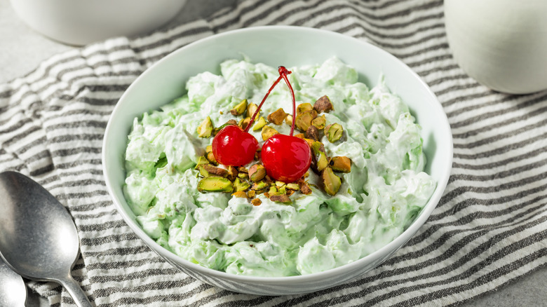 a bowl of watergate salad topped with pistachios and two red cherries on a gray and white striped placemat