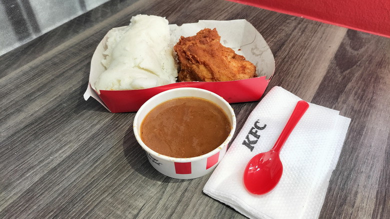 A container of KFC gravy next to a paper tray with chicken and mashed potatoes