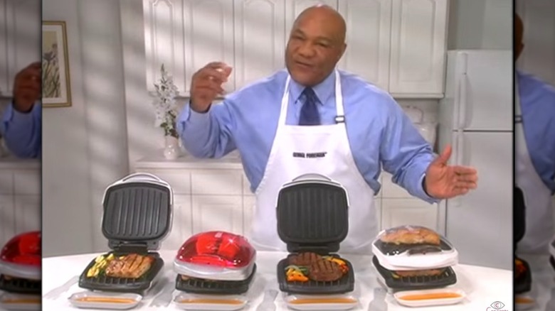 George Foreman wearing an apron standing behind four George Foreman grills in an infomercial