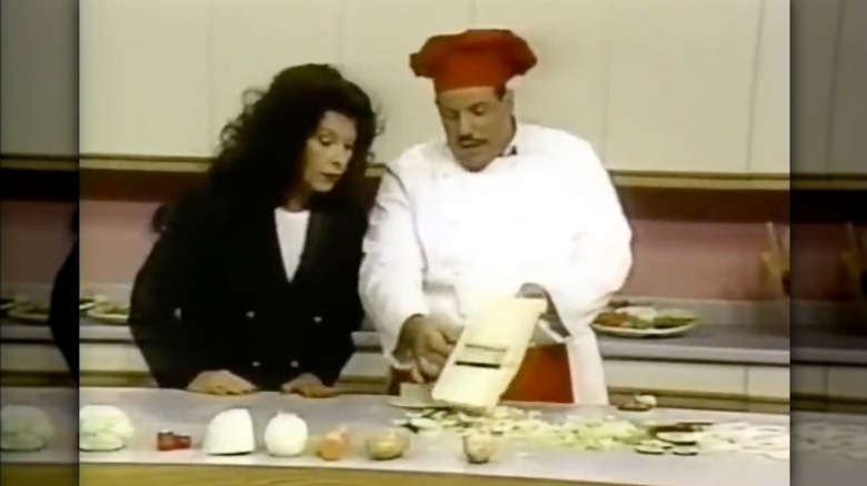 Man in a chef's coat and red chef's hat slicing an onion on a Kitchen Wizard Plus while a woman in a black blazer watches