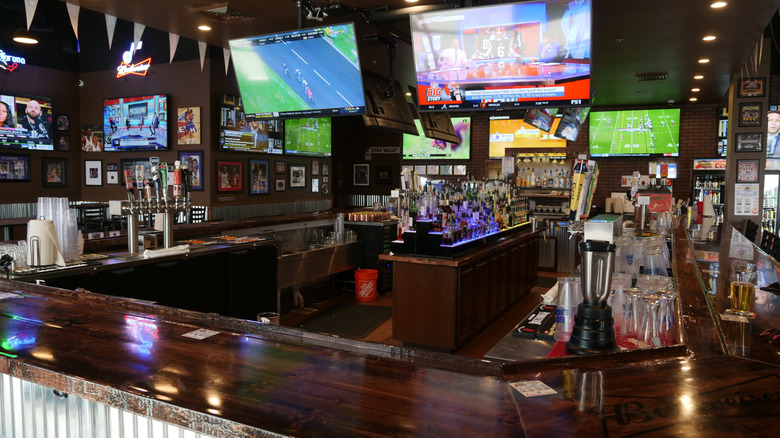An image of an empty-looking sports bar features a dozen or so television screens showing sports coverage, along with beer taps, alcohol bottles, and a blender.