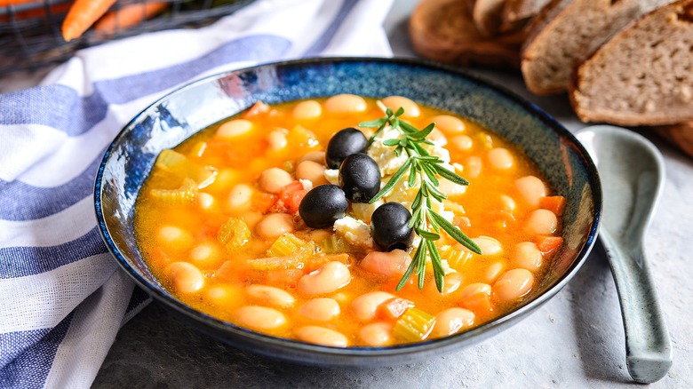 Greek white bean soup in a blue-patterned bowl topped with whole black olives and a sprig of fresh rosemary