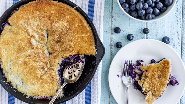 blueberry cobbler in a cast iron pan with a slice on a whie plate beside it