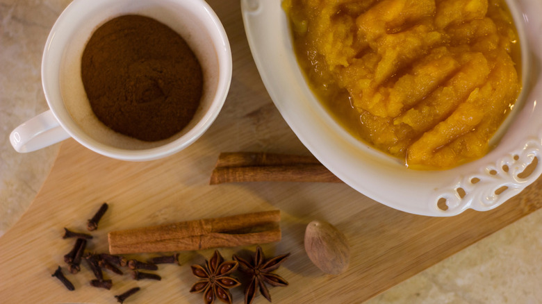 Pumpkin purée in dish next to cinnamon in tea cup with other whole spices on cutting board.