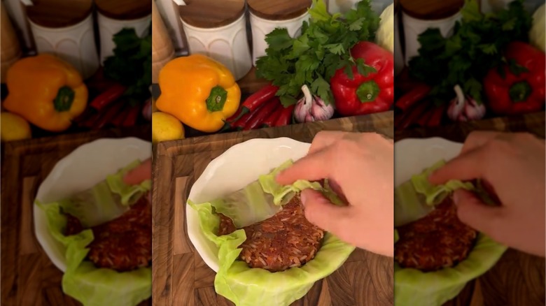 A hand rolling cabbage leaves filled with stuffing in a white bowl, with fresh produce in the background.