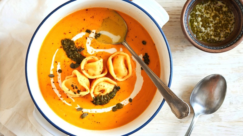 Creamy tortellini soup with pumpkin puree and pesto in a white bowl with blue trim and two spoons.