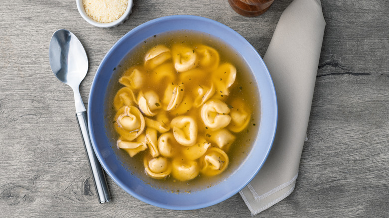 Broth-based tortellini soup in a blue bowl with spoon and napkin.