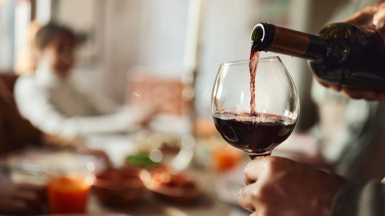 Hand pouring red wine into a glass with a person sitting in the distance