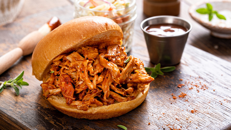 Pulled chicken sandwich on a cutting board with herbs, seasoning, and a small cup of BBQ sauce