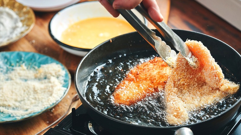 Person using metal tongs to flip panko-encrusted chicken filets in hot oil in a frying pan