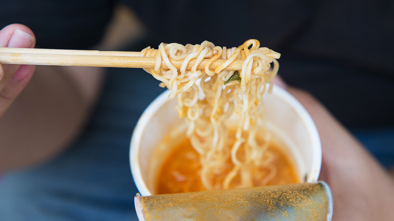Person's hand holding chopsticks of instant noodles.