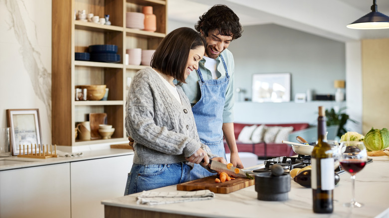 Two people are seen smiling and cooking together in a kitchen; an open bottle of wine is nearby.