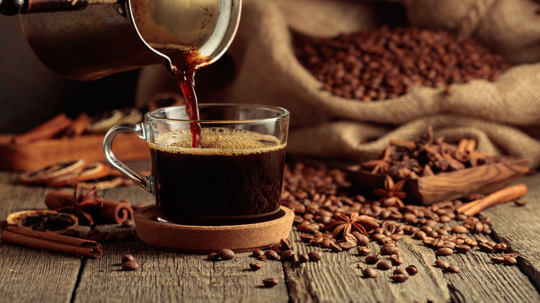 A freshly brewed cup of coffee is served from a pot with coffee beans, cinnamon, and other spices on a table around the cup