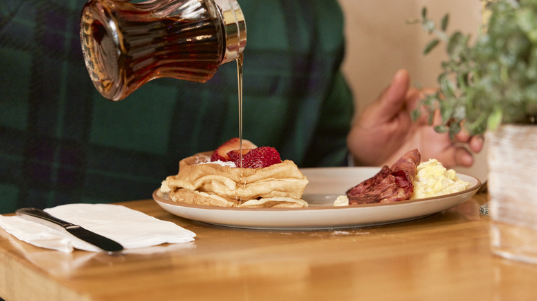 Maple syrup being drizzled over a stack of pancakes topped with strawberries, with bacon and eggs on the side