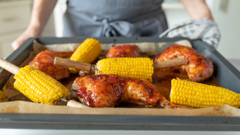 Person taking roasted chicken and corn on the cob on a sheet pan out of the oven