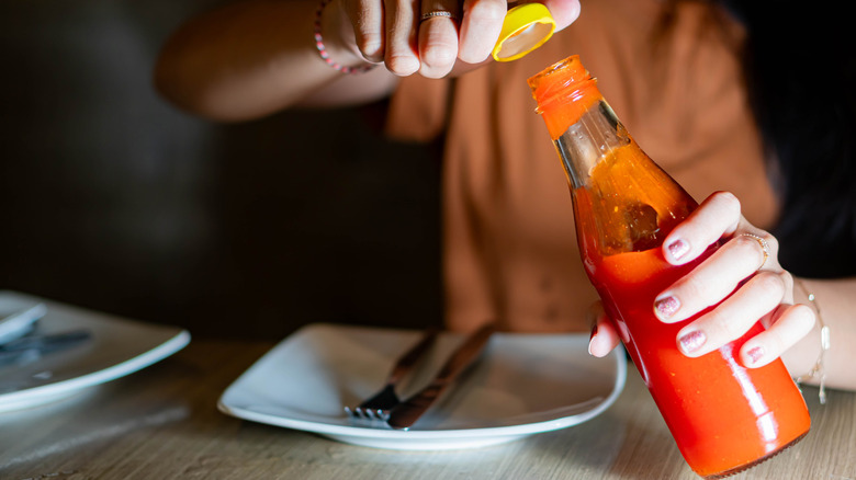 person opening an unlabeled hot sauce bottle with a place setting in front of them