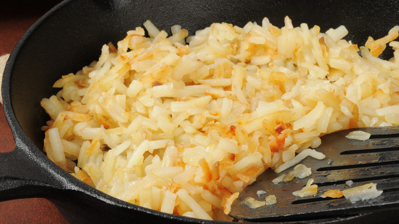 Partially cooked hash browns in a cast iron pan with spatula in corner.