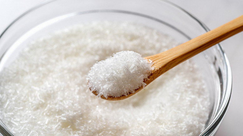 Monosodium glutamate in a wooden spoon over glass bowl full of MSG white crystals close-up