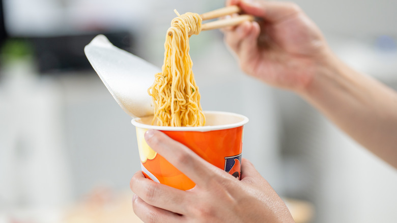 Hands holding chopsticks, eating instant noodles in paper cup at home