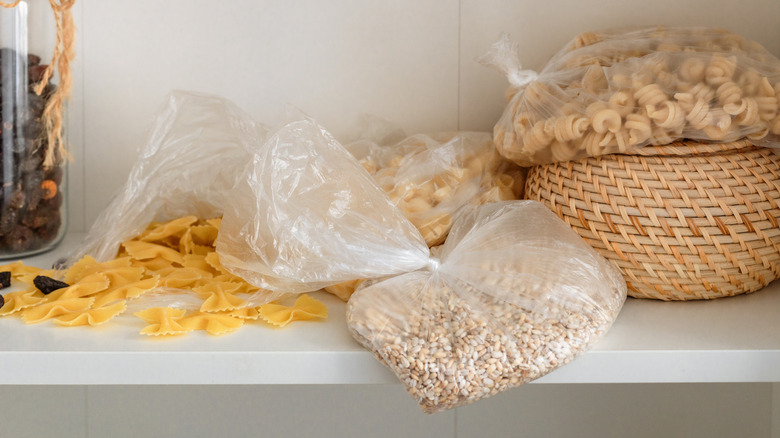 A messy kitchen shelf with pasta and oats in plastic, with some pasta spilling out of its container.