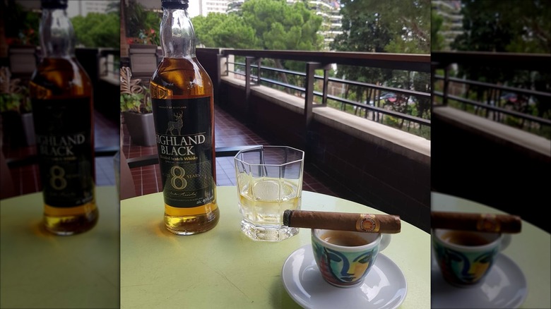A bottle of Aldi's Highland Black scotch on an apartment balcony table next to a rocks glass filled with ice and whisky, and a cigar balanced over a cup of coffee