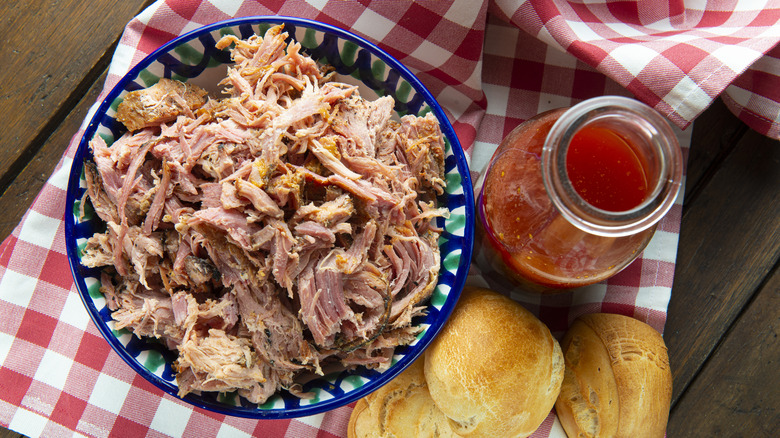 A bowl of pulled pork with a bottle of barbecue sauce and rolls