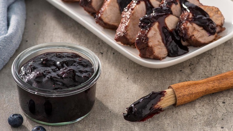 A jar of blueberry barbecue sauce next to a brush and plate of sliced pork
