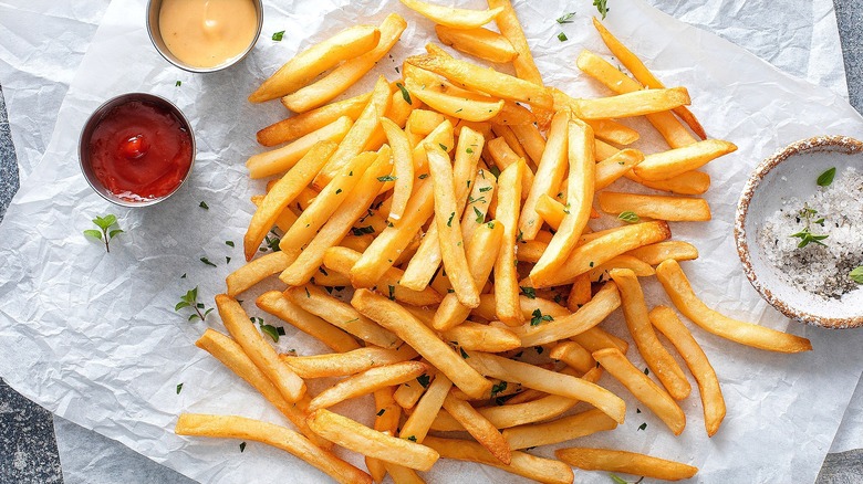 French fries piled on two sheets on white parchment paper surrounded by small bowls of condiments and salt