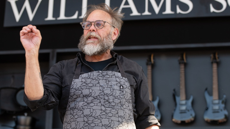 Alton Brown at a cooking demonstration, wearing a grey apron in front of a line of guitars