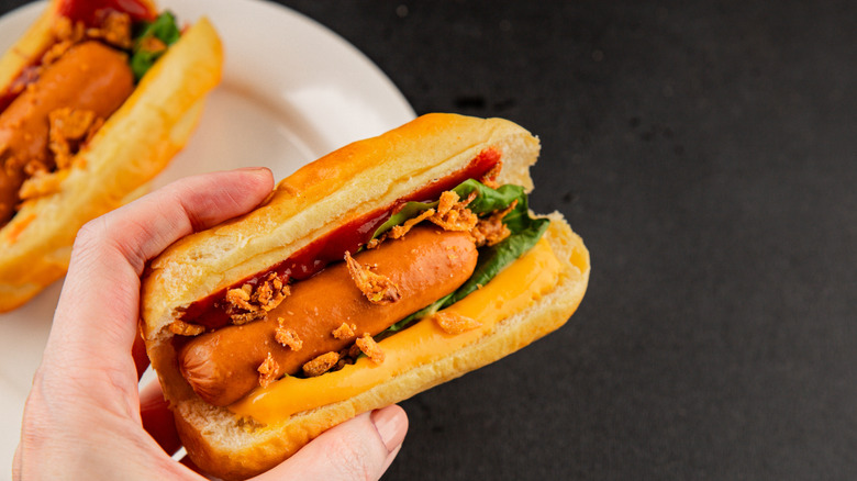 Close up of hand holding hotdog bun with hotdog, condiments, and fried onions and lettuce