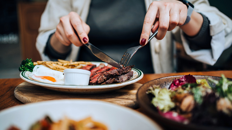 Two hands cutting a grilled steak with french fries and a side salad