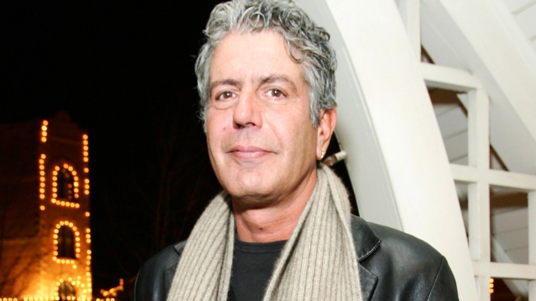 Anthony Bourdain in a black leather jacket and beige scarf posing in front of a building.