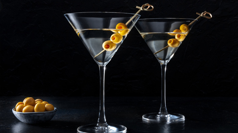 Two dry martinis in martini glasses with three olives on a bamboo swizzle stick, a small bowl of olives to the side.