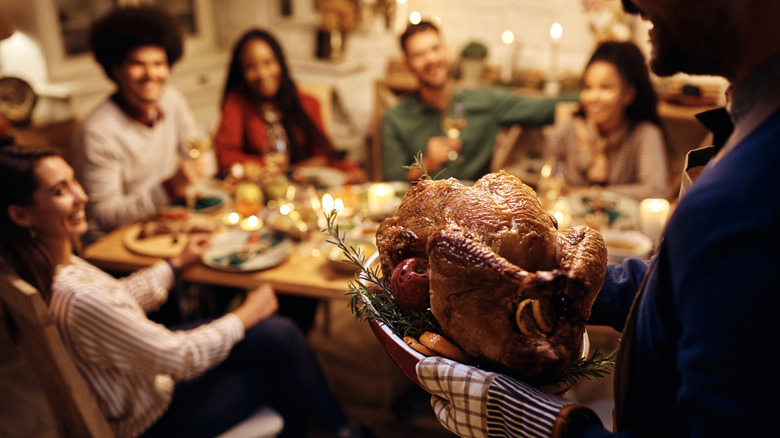 Man carrying a turkey to a Thanksgiving table surrounded by people