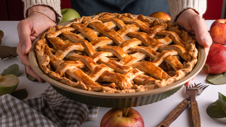 Hands holding a homemade apple pie over a table with apples and silverware.