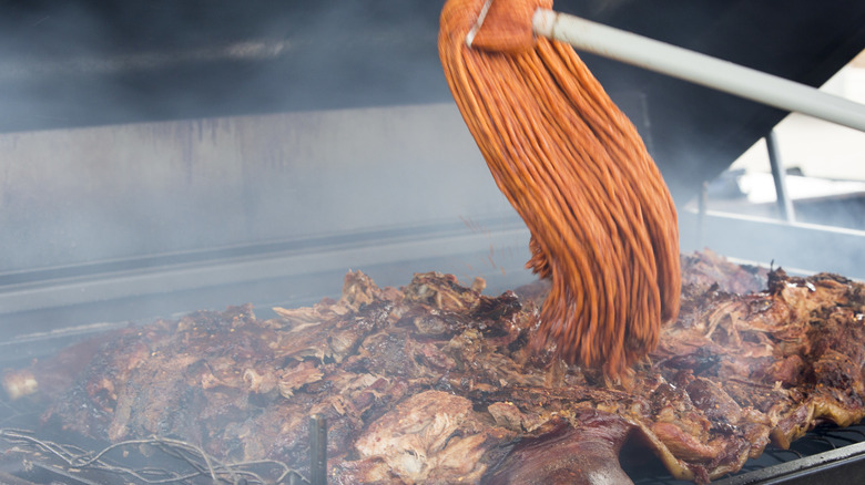 An individual using a barbecue mop to spread sauce over meat on a hot grill.