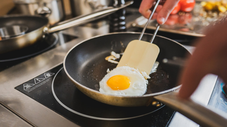 A close-up of a chef's hand using a spatula to flip a sunny-side-up egg in a frying pan.