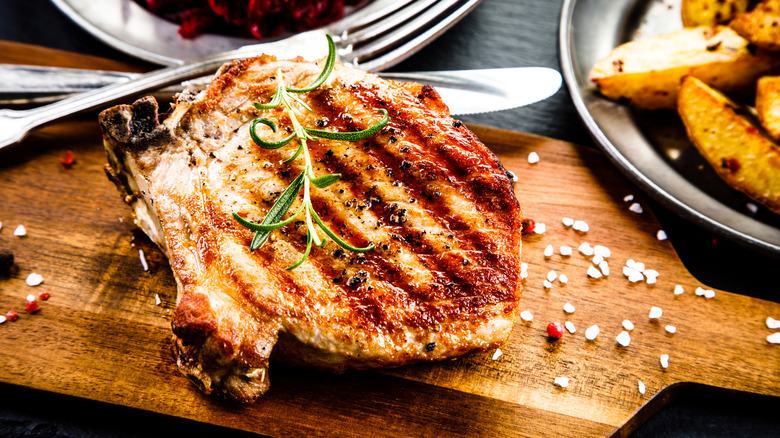 A grilled pork chop on a wooden board with an herb garnish and chunky salt on the board.