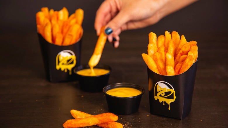 Hand dipping a Taco Bell nacho fry into nacho cheese, along with two containers of nacho fries