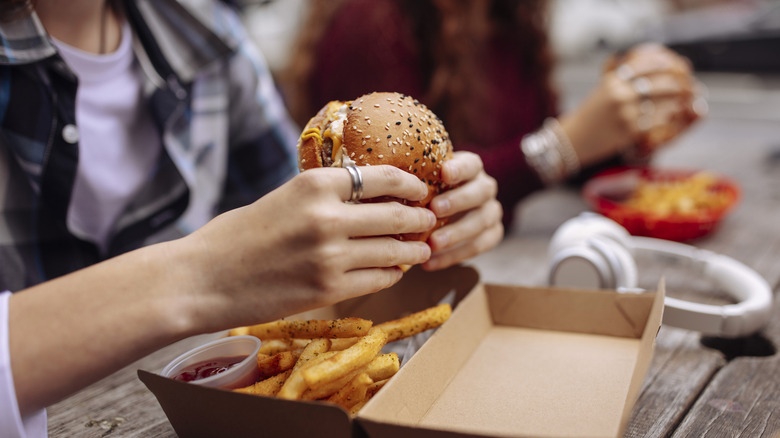 Close up of someone holding burger at street food restaurant while sitting outdoors at the table with his friends