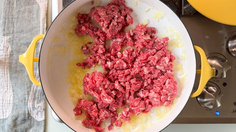 Raw cubed flatiron steak in yellow pot with diced yellow onion on stovetop