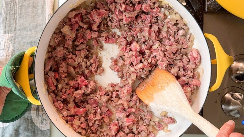 Stirring cubed flatiron steak and diced onion in large yellow pot on stovetop with wooden spoon